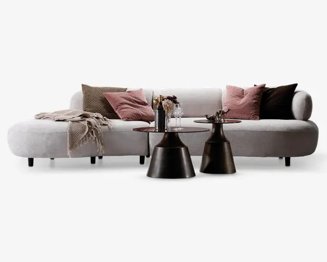 Moderne L-formet sofa betrukket med lysegråt stof, med lav ryg og afrundede kanter. Sofaen er pyntet med forskellige pyntepuder i nuancer af rosa, brun og beige samt et beige plaid med frynser, der er lagt over den ene arm. Foran sofaen står to kegleformede sideborde i mørk bronzefarvet metal, hvorpå der står dekorationer som en vinflaske, glas og små vaser med blomster.