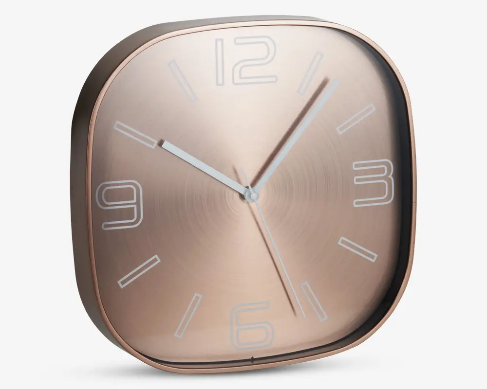 Moderne vægur med afrundet firkantet form og metallisk rosaguld finish. Uret har minimalistiske hvide time-markeringer samt tal ved 12, 3, 6 og 9. De tre slanke visere til timer, minutter og sekunder er i hvid, hvilket giver god læsbarhed.