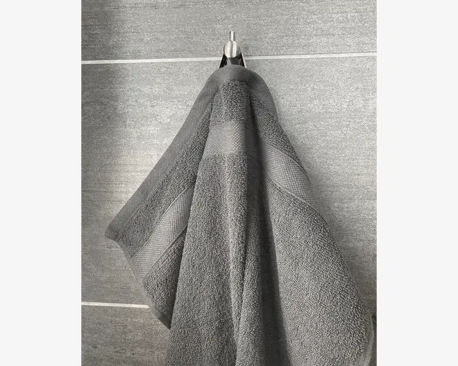 Et mellemstort, mørkegråt håndklæde hænger på en metalkrog mod en grå flisevæg. Håndklædet har en tekstureret, absorberende overflade med en diskret vævet kant og et dekorativt bånd, der løber vandret nær kanten. Stoffet ser tykt og blødt ud, velegnet til at tørre sig efter bad eller brusebad.