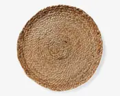 Rund dækkeserviet fremstillet af tæt flettede naturfibre fra søgræs, med en rustik, tekstureret overflade og synlig fletning i et spiralformet mønster. Kanterne er let hævede, hvilket giver et robust og håndlavet udtryk. Farven er en blanding af lyse og mellembrune nuancer, der fremhæver det organiske materiale.