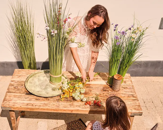 Et rustikt træbord står udendørs med et stort grønt keramikfad, en rulle snor og en buket kunstige vilde blomster og bær. Høje, dekorative græsarrangementer med små farverige blomster står i vaser på og omkring bordet. Scenen er lys og solrig og antyder en kreativ eller blomsterdekorationsaktivitet.