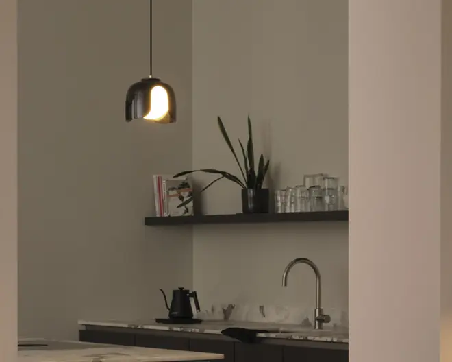 Et moderne køkken med en mat sort pendellampe, der har et afrundet design og varm belysning. Bordpladen er lys med diskrete årer, og skabene nedenunder er mørke. En elegant, buet vandhane i rustfrit stål er monteret over en dyb vask. På væggen er der en svævende hylde med en potteplante med lange, spidse blade, flere stablede klare glas og et par kogebøger. En sort elkedel står på bordet og bidrager til det minimalistiske og moderne udtryk.