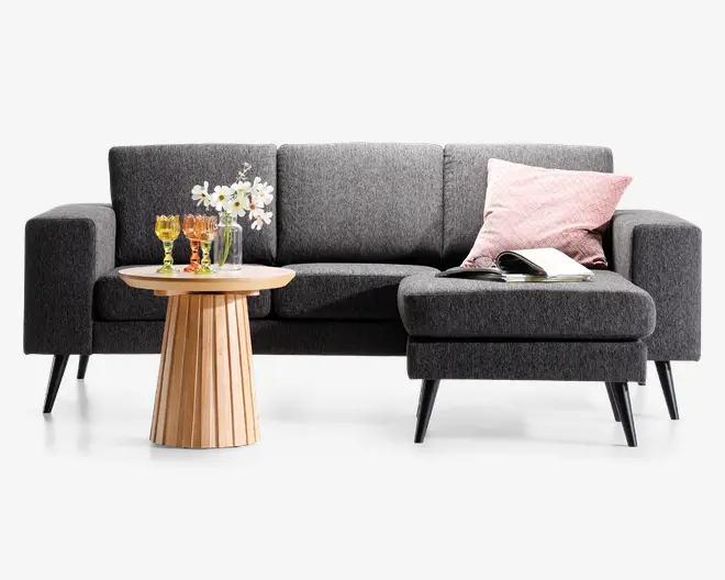 Moderne L-formet sofa betrukket med mørkegråt stof, rene linjer og slanke, skrå sorte ben. Sofaen har en chaiselong på højre side, en lyserød pude med tekstur og et åbent magasin liggende på sædet. Foran sofaen står et rundt sofabord i træ med rillet fod, hvorpå der står tre farvede glas og en lille vase med hvide blomster.