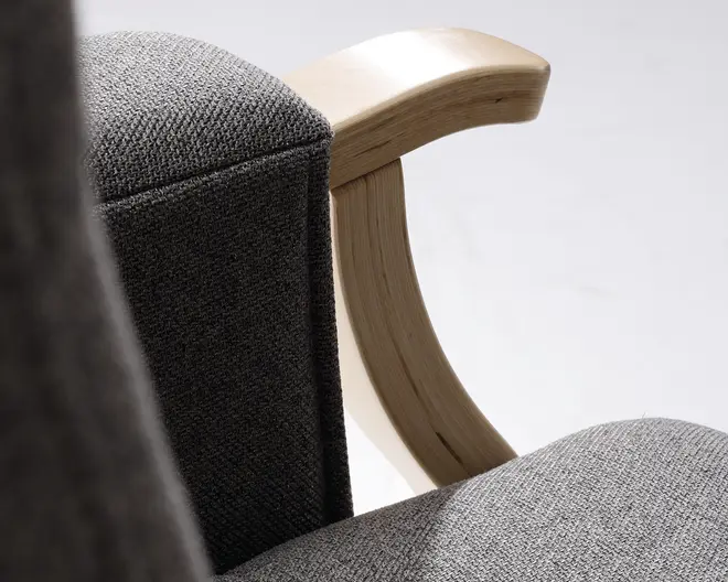 Nærbillede af en stol med buet armlæn i lyst træ og polstring i gråt, tekstureret stof. Stoffet har en tæt vævning, hvilket giver et slidstærkt og moderne udtryk. Armlænet er glat og velpoleret, hvilket står i kontrast til det bløde, polstrede sæde og ryglæn.