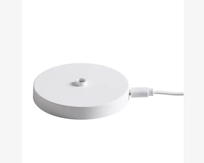 En rund, flad, hvid base med et lille centralt hul og en tilsluttet hvid strømkabel. Overfladen er glat og minimalistisk, og kablet er forbundet til siden af basen via et USB-stik.