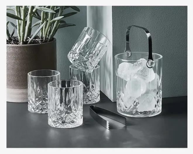 Sæt med fire klare, facetslebne glas med klassisk slibemønster, ledsaget af en matchende isspand fyldt med isterninger. Isspanden har et solidt metalhåndtag og leveres med en tang i metal. Glassene og spanden har en tung, elegant bund og en skinnende, tekstureret overflade, der reflekterer lyset.