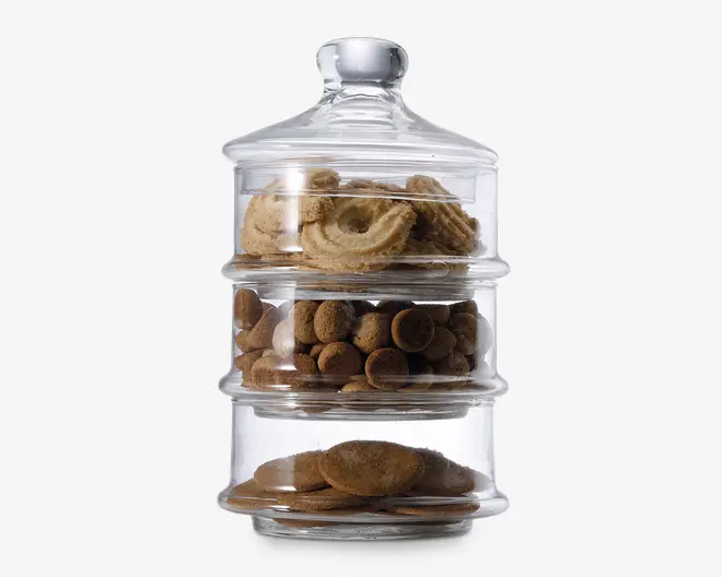 Klar, stabelbar glasbeholder med tre separate rum, hver fyldt med forskellige slags småkager. Øverste rum indeholder runde, snørklede smørsmåkager, midterste rum har små, runde chokoladekiks, og nederste rum har flade, ovale småkager. Beholderen har et tætsluttende låg med knop, som sikrer lufttæt opbevaring og holder indholdet frisk.