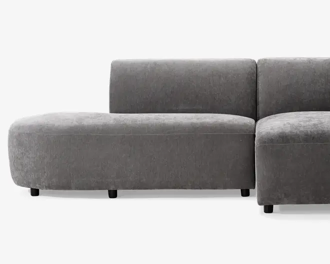 Moderne modulsofa med blødt, plysset gråt stofbetræk. Sofaen har en afrundet venstre armlæn og ryglæn, hvilket giver et glat og nutidigt udtryk. Sædehynderne er tykke og støttende, og sofaen står på korte, solide sorte ben. Designet er minimalistisk med rene linjer og uden synlige sømme eller udsmykninger.