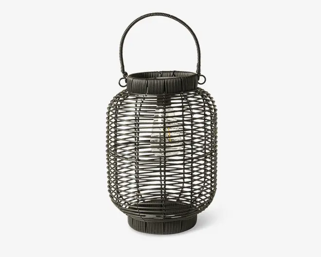 Mørkfarvet lanterne med flettet rattan-ydre, cylindrisk form der smalner let ved top og bund. Lanternen har et solidt håndtag fastgjort øverst, hvilket gør den nem at bære eller hænge op. Indvendigt ses en LED-pære omsluttet af et gennemsigtigt dæksel, hvilket giver et moderne præg til det rustikke design. Den åbne fletning lader lyset skinne igennem og skaber et dekorativt mønster, når lanternen er tændt.