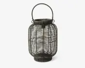 Mørkfarvet lanterne med flettet rattan-ydre, cylindrisk form der smalner let ved top og bund. Lanternen har et solidt håndtag fastgjort øverst, hvilket gør den nem at bære eller hænge op. Indvendigt ses en LED-pære omsluttet af et gennemsigtigt dæksel, hvilket giver et moderne præg til det rustikke design. Den åbne fletning lader lyset skinne igennem og skaber et dekorativt mønster, når lanternen er tændt.