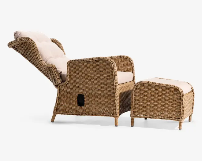 Liggestol med tilhørende fodskammel, begge med flettet rattan-design i en naturlig lys brun farve. Stolen har høj ryg og armlæn samt tykke, lyse beige hynder for ekstra komfort. Liggemekanismen er synlig på siden, så ryglænet kan lænes bagud for en mere afslappet siddeposition. Fodskamlen har også en polstret top, der giver en behagelig støtte til fødderne.