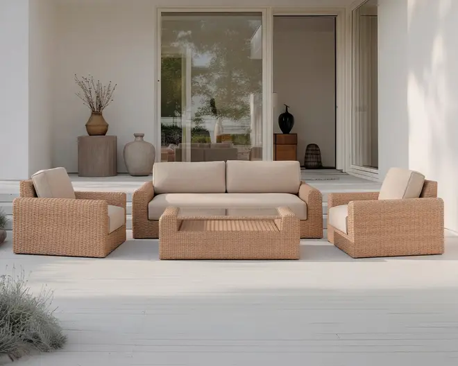 Udendørs loungesæt bestående af en trepersoners sofa, to lænestole og et rektangulært sofabord, alle med flettet rattan-lignende stel i en naturlig lysebrun farve. Hynderne er tykke og betrukket med et lyst beige stof, hvilket giver et komfortabelt og indbydende udtryk. Designet er moderne og minimalistisk, velegnet til terrasser eller haveområder.