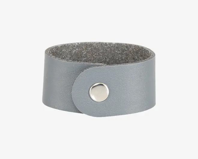 Bred grå lædermanchet med en glat, mat overflade og en enkelt lukning med sølvfarvet trykknap. Indersiden er foret med et blødt, tekstureret materiale for komfort.