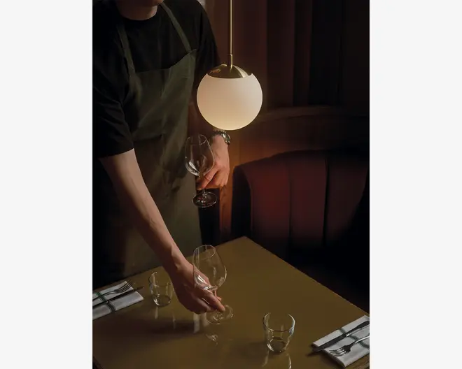 En person iført mørk skjorte og olivengrøn forklæde dækker et bord under varm, stemningsfuld belysning. Bordet har en blank overflade og er dækket med klare vinglas, små vandglas og pænt foldede servietter med bestik. En moderne, kugleformet pendellampe hænger over bordet og kaster et blødt lys over scenen. I baggrunden ses en polstret bås i afdæmpede farver, hvilket skaber en hyggelig og elegant spiseatmosfære.