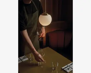 En person iført mørk skjorte og olivengrøn forklæde dækker et bord under varm, stemningsfuld belysning. Bordet har en blank overflade og er dækket med klare vinglas, små vandglas og pænt foldede servietter med bestik. En moderne, kugleformet pendellampe hænger over bordet og kaster et blødt lys over scenen. I baggrunden ses en polstret bås i afdæmpede farver, hvilket skaber en hyggelig og elegant spiseatmosfære.