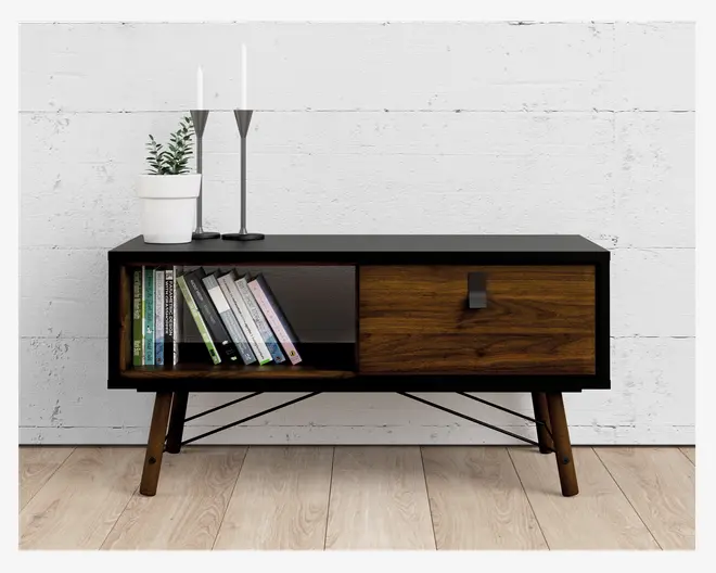 Rektangulært skab i træ med sort top og ramme, skråtstillede træben og metalstøtter underneden. Venstre side har en åben hylde med flere bøger, mens højre side har en skydedør med mørkt håndtag og synlig træstruktur. Ovenpå står en hvid urtepotte med grøn plante samt to høje, slanke sølvlysestager med hvide stearinlys.