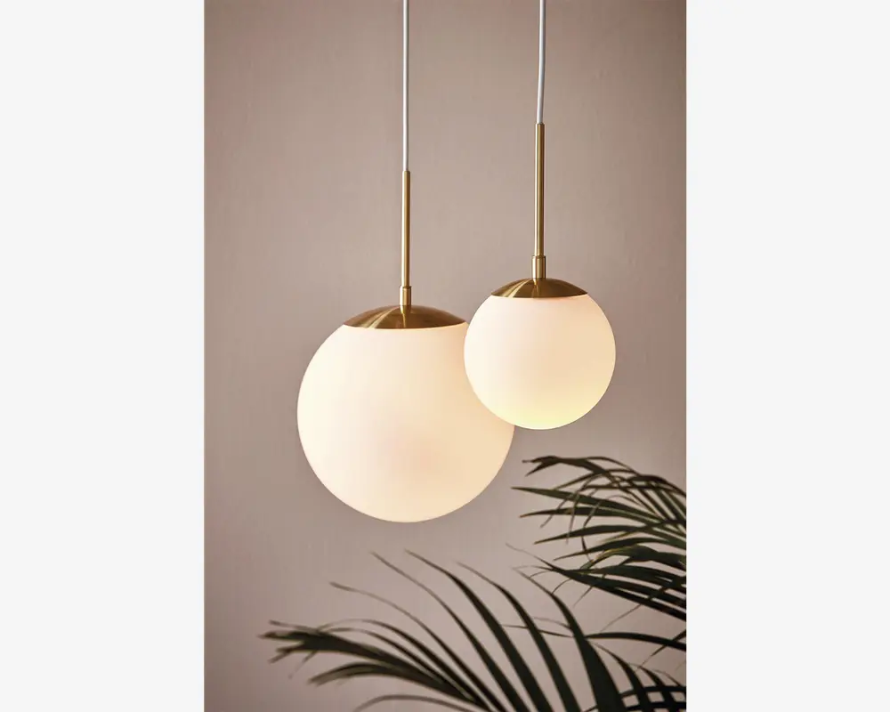 To pendellamper med kugleformede, matthvide glasskærme og detaljer i børstet messing. Den ene kugle er større og hænger lidt lavere end den mindre. Begge lamper er ophængt i hvide ledninger. Designet er moderne og elegant og giver et blødt, varmt lys. En bladplante ses delvist nederst i billedet.