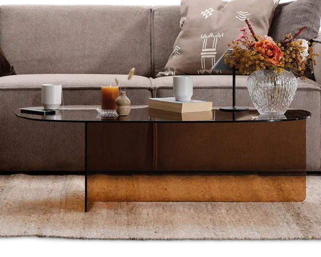 Ovalt sofabord med en blank, mørktonet glasplade og en reflekterende bronzefarvet base. Bordet har et minimalistisk design med tynde, sorte metalben. På bordpladen står der dekorative genstande, herunder et hvidt krus, et brunt stearinlys, en lille beige vase med et siv, to stablede bøger, en sort lampe og en klar glasvase med en buket orange og gule blomster. Bordet står på et lyst, tekstureret tæppe foran en sofa i neutrale farver med mønstrede puder.