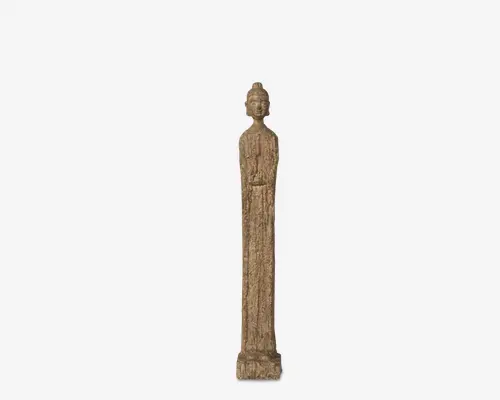Høj, slank statue med en tekstureret, stenlignende overflade. Figuren står oprejst med armene foldet foran sig og bærer en lang, flydende kappe, der går helt ned til soklen. Hovedet har en topknude, og det samlede design er minimalistisk med diskrete detaljer på klædningen. Ansigtet er bevidst sløret.