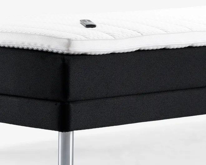 Nærbillede af et sengehjørne med en tyk, struktureret hvid topmadras, der har en synlig lynlås og et lille mærke. Sengebunden er betrukket med sort stof med en diskret vævet struktur, og sengen står på et slankt, cylindrisk metalben.