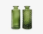 To grønne glasflasker med brede, flade åbninger og cylindrisk form. Flasken til venstre har lodrette rillede linjer, der giver en struktureret overflade. Flasken til højre har et diamantmønster, som giver et mere detaljeret og dekorativt udtryk. Begge flasker er gennemsigtige og har et vintage-inspireret design.
