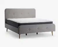 En moderne sengegavl betrukket med lysegråt stof, med en høj, afrundet hovedgavl og en matchende fodgavl, begge med knapdetaljer. Sengen står på fire skråtstillede træben med naturfinish. Madrassen er af medium tykkelse med hvid, quiltet top og mørkegrå sider.