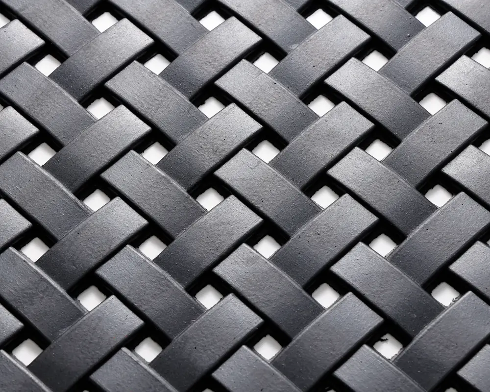 Nærbillede af et tæt vævet mønster lavet af flade, mat sorte strimler. Strimlerne er flettet sammen i et over-under kurveflet-design, hvilket skaber en tekstureret og holdbar overflade. Materialet virker robust, og vævningen er ensartet, hvilket giver både styrke og et moderne udtryk.