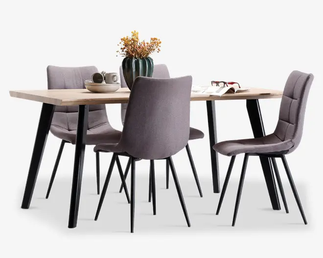 Sættet består af et rektangulært bord i lyst træ med sorte skrå ben og fire polstrede stole i en afdæmpet grå farve. Stolene har et moderne, polstret design med diskrete horisontale syninger og sorte metalben. Bordet er dekoreret med en vase med tørrede blomster, en skål, kopper og en åben bog, hvilket fremhæver den rummelige bordplade og det moderne udtryk.