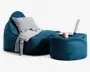 Et moderne loungemiljø med en dyb teal-farvet, tekstureret sækkestol og en matchende puf. Sækkestolen har en blød, grå pude og et lysegråt tæppe lagt over den ene side, hvilket giver ekstra komfort og stil. Puffen fungerer som et lille bord og har en bærbar computer, et lyserødt krus og en notesbog ovenpå, hvilket viser dens alsidighed til både afslapning og arbejde. Stoffet ser slidstærkt ud, og det samlede design er afslappet og moderne.