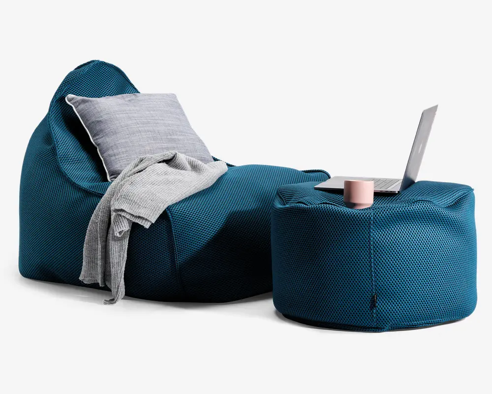 Et moderne loungemiljø med en dyb teal-farvet, tekstureret sækkestol og en matchende puf. Sækkestolen har en blød, grå pude og et lysegråt tæppe lagt over den ene side, hvilket giver ekstra komfort og stil. Puffen fungerer som et lille bord og har en bærbar computer, et lyserødt krus og en notesbog ovenpå, hvilket viser dens alsidighed til både afslapning og arbejde. Stoffet ser slidstærkt ud, og det samlede design er afslappet og moderne.