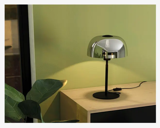 En moderne bordlampe med en blank, metallisk kuppelformet skærm og en slank sort stander. Lampen har en rund, flad fod og en synlig sort ledning med en afbryder på ledningen. Den reflekterende overflade på skærmen giver lampen et stilrent og moderne udtryk, og lampen kaster et varmt, fokuseret lys på væggen og overfladen nedenunder.