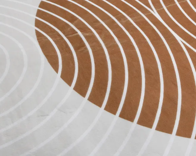 Nærbillede af en stofoverflade med et moderne geometrisk design. Mønstret består af buede hvide linjer, der danner koncentriske buer over en baggrund, som skifter fra lys beige til en varm brun sektion. Teksturen ser blød og glat ud, hvilket indikerer et behageligt materiale.