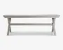 Rektangulært bord med en overflade af lameller, udført i en lys grå farve. Bordet har solide X-formede ben i hver ende, forbundet af en vandret støttebjælke for ekstra stabilitet. Designet er minimalistisk og moderne, velegnet til både indendørs og udendørs brug.
