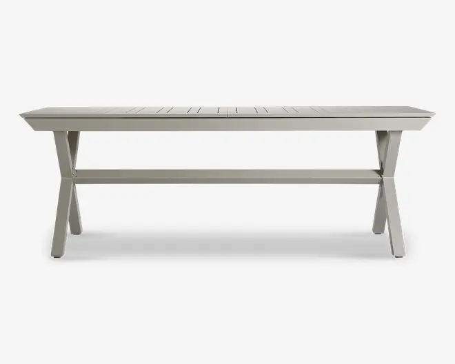 Rektangulært bord med en overflade af lameller, udført i en lys grå farve. Bordet har solide X-formede ben i hver ende, forbundet af en vandret støttebjælke for ekstra stabilitet. Designet er minimalistisk og moderne, velegnet til både indendørs og udendørs brug.