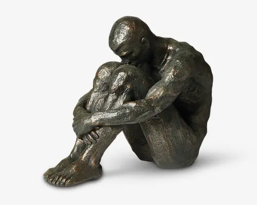 Bronzefarvet skulptur af en siddende mandlig figur med en tekstureret overflade. Figuren sidder med knæene trukket op til brystet, armene omkring benene og hovedet bøjet fremad i en eftertænksom eller indadvendt positur. Detaljerne fremhæver muskelstruktur og realistiske proportioner, med synlige fingre og tæer. Overfladen har diskrete højdepunkter, der fremhæver form og tekstur.