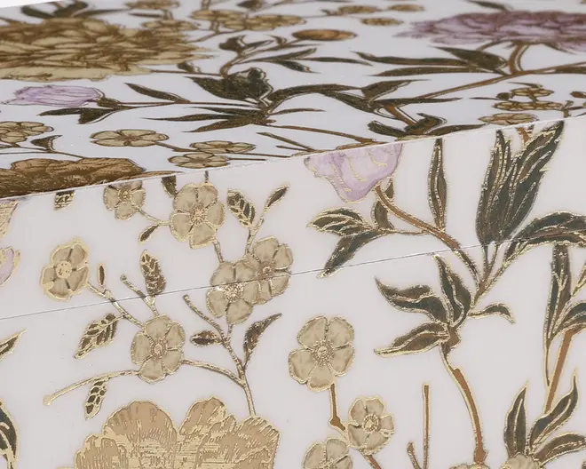 Nærbillede af en overflade med et indviklet blomstermønster i guld, grøn og lyse lilla nuancer. Blomsterne og bladene er præget, hvilket giver en tekstureret, forhøjet effekt. Baggrunden er hvid, hvilket får de metalliske og pastelfarver til at træde tydeligt frem. Designet fremstår elegant og luksuriøst, velegnet til dekoration eller gaveformål.