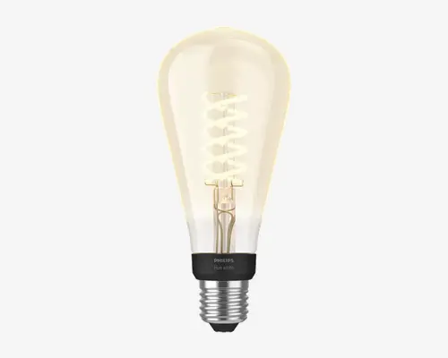 En LED-pære i retrostil med klart, dråbeformet glas og en synlig spiralformet glødetråd, der udsender et varmt hvidt lys. Soklen er i metal med en sort krave, hvor der står 'PHILIPS Hue White.' Pæren forener et klassisk udseende med moderne smart belysningsteknologi.