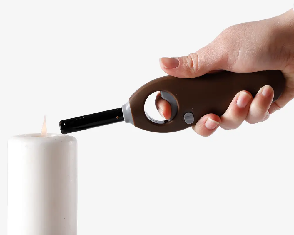 En hånd holder en brun lang lighter med sølvfarvet tændingsknap og ergonomisk buet håndtag, som bruges til at tænde et hvidt bloklys. Lighteren har en sikkerhedsring til fingerplacering og et sort mundstykke for præcis flammekontrol.