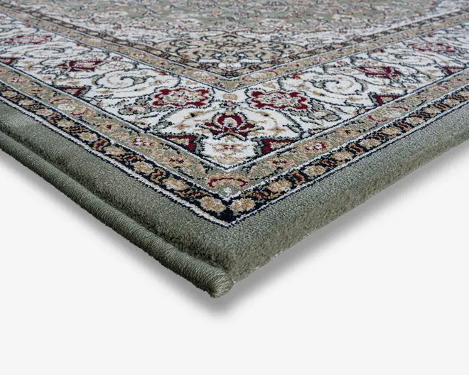 Nærbillede af et tæppehjørne, der viser en tyk, blød luv og tæt indbundne kanter. Tæppet har et indviklet, traditionelt mønster med blomster- og geometriske motiver i nuancer af creme, rød, grøn og beige. Kanten er detaljeret med gentagne designs, og overfladen ser blød og tæt ud, hvilket indikerer holdbarhed og komfort.