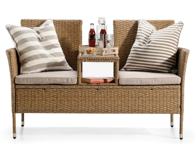 To-personers bænk lavet af flettet rattan med indbygget centertable. Bænken har to tykke, lyse sædehynder og to store pyntepuder med grå og hvide striber. Centertablet har en flad overflade med drikkevarer og snacks samt en åben hylde nedenunder, hvor der ligger en bog. Benene er slanke og har metalforstærkede fødder for ekstra stabilitet.