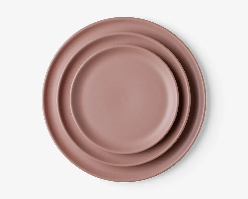 Sæt med tre runde tallerkener i en afdæmpet rosa farve, hver tallerken lidt større end den næste, med en glat, mat overflade og et minimalistisk design. Tallerkenerne har en let hævet kant og ingen synlige mønstre eller dekorationer.