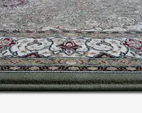 Nærbillede af et tykt, blødt tæppe med detaljeret blomster- og snirklet mønster. Tæppet har en grøn bundfarve med creme, beige og bordeauxrøde detaljer. Kanten har indviklede designs med blandede farver, og luven ser tæt og blød ud, hvilket indikerer en behagelig tekstur.