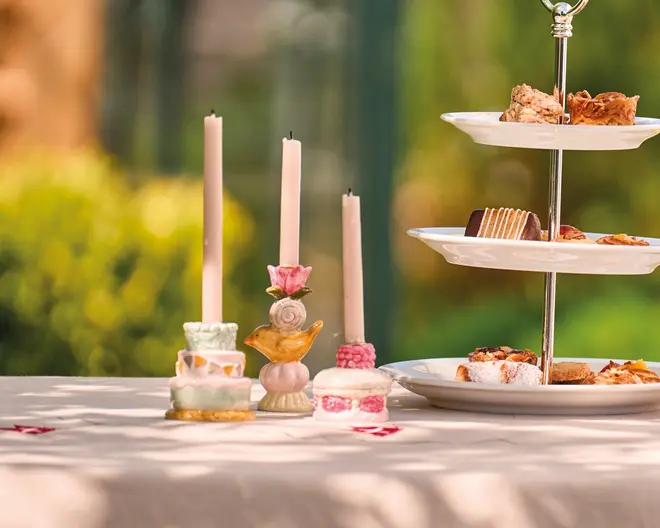 Billedet viser et bord dækket til en udendørs sammenkomst med tre dekorative lysestager i pastelfarver og blomsterdesign, hver med et højt, slankt stearinlys. Ved siden af står et etagestativ med to niveauer, fyldt med forskellige kager og søde sager, herunder lagkager og småkager. Dugens farve er lys, og baggrunden er sløret med grønne planter, hvilket antyder en have.