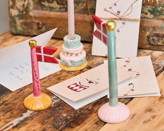 Billedet viser en samling dekorative genstande på en rustik træoverflade. Der er to små keramiske flagstænger, hver med et dansk flag og en guldtoppet knop, den ene med gul base og teksten 'TILLYKKE' skrevet på stangen, og den anden med lyserød base. Der ses også en pastelfarvet keramisk lysestage formet som en lagkage. Flere lykønskningskort ligger spredt omkring, nogle med illustrationer af danske flag og festlige motiver, og et kort er bundet med snor. Scenen har et festligt og håndlavet udtryk.