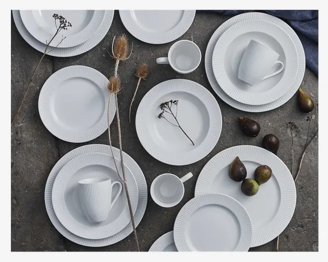 Et sæt hvidt keramisk stel med tallerkener, skåle og kopper, der har en diskret lodret rillet struktur langs kanterne. Designet er minimalistisk og elegant med rene linjer og blank overflade. Sættet indeholder forskellige størrelser af tallerkener og kopper, velegnet til forskellige måltider.