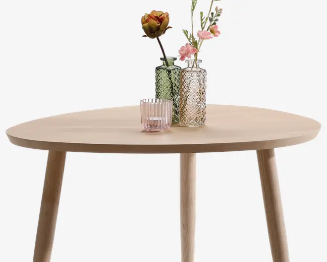 Rundt bord i lyst træ med glat overflade og tre skrå ben. Ovenpå står tre dekorative glasvaser i grønne og lyserøde nuancer, hver med sit eget mønster i glasset. To af vaserne indeholder kunstige blomster i gule, lyserøde og hvide farver, som giver et farverigt og elegant udtryk.