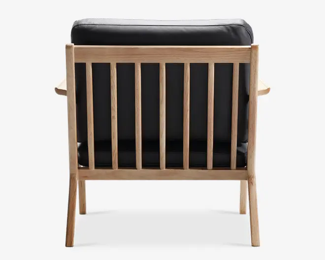 Billedet viser bagsiden af en stol med en ramme af lyst træ og lodrette lameller. Stolen har en sort polstret ryg og sæde, hvilket giver en kontrast til det lyse træ. Benene er let skråtstillede, og armlænene, som også er lavet af træ, er synlige på begge sider. Designet er minimalistisk og moderne, og fremhæver både træets håndværk og komforten fra de polstrede puder.