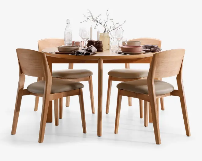 Sættet består af et rundt bord i lyst træ med glat overflade og fire matchende stole med buede rygge og polstrede beige sæder. Bordet er dækket med moderne service, herunder tallerkener, skåle, glas og servietter samt dekorative elementer som en vase med grene, stearinlys og en vandkaraffel. Designet er minimalistisk og moderne, velegnet til en spisestue.