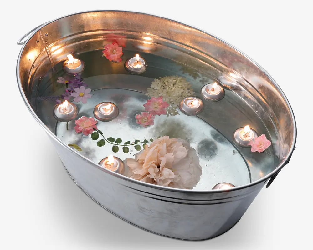 En stor, ovalformet metalkar fyldt med vand. Flere små fyrfadslys flyder på overfladen, alle tændte og udsender et varmt skær. Der er også forskellige kunstige eller ægte blomster i bløde pastelfarver som lyserød, hvid og lilla, der flyder mellem lysene. Karret har en blank, reflekterende overflade og en glat kant, hvilket giver det et rent og elegant udseende.