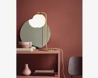 En moderne bordlampe med en rund, mat hvid glasskærm og en slank, guldfarvet metalramme. Lampen har en buet arm og en flad, cirkulær fod. Den står på et støvet lyserødt konsolbord ved siden af et rundt spejl, stablede bøger og en lille lyserød vase. Lampens ledning er synlig og hænger ud over siden af bordet. Bagvæggen er malet i en varm, rødbrun farve.